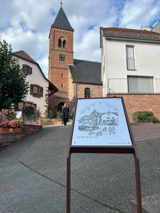 Panneau visuel sur pied dans une rue de Dossenheim-sur-Zinsel, église en arrière-plan